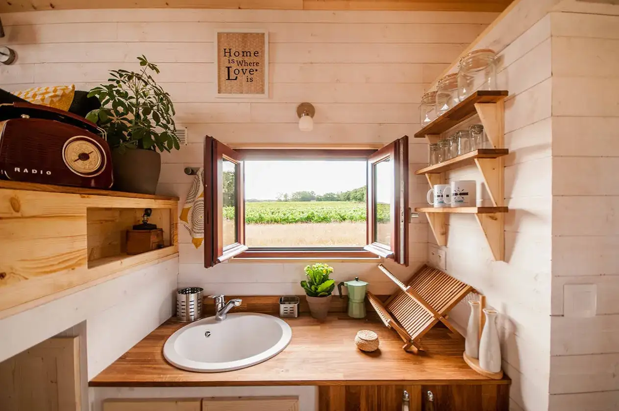 Intérieur d'une maison sur roulette avec un évier, une cafetière verte et une vue sur un champ verdoyant par la fenêtre ouverte.