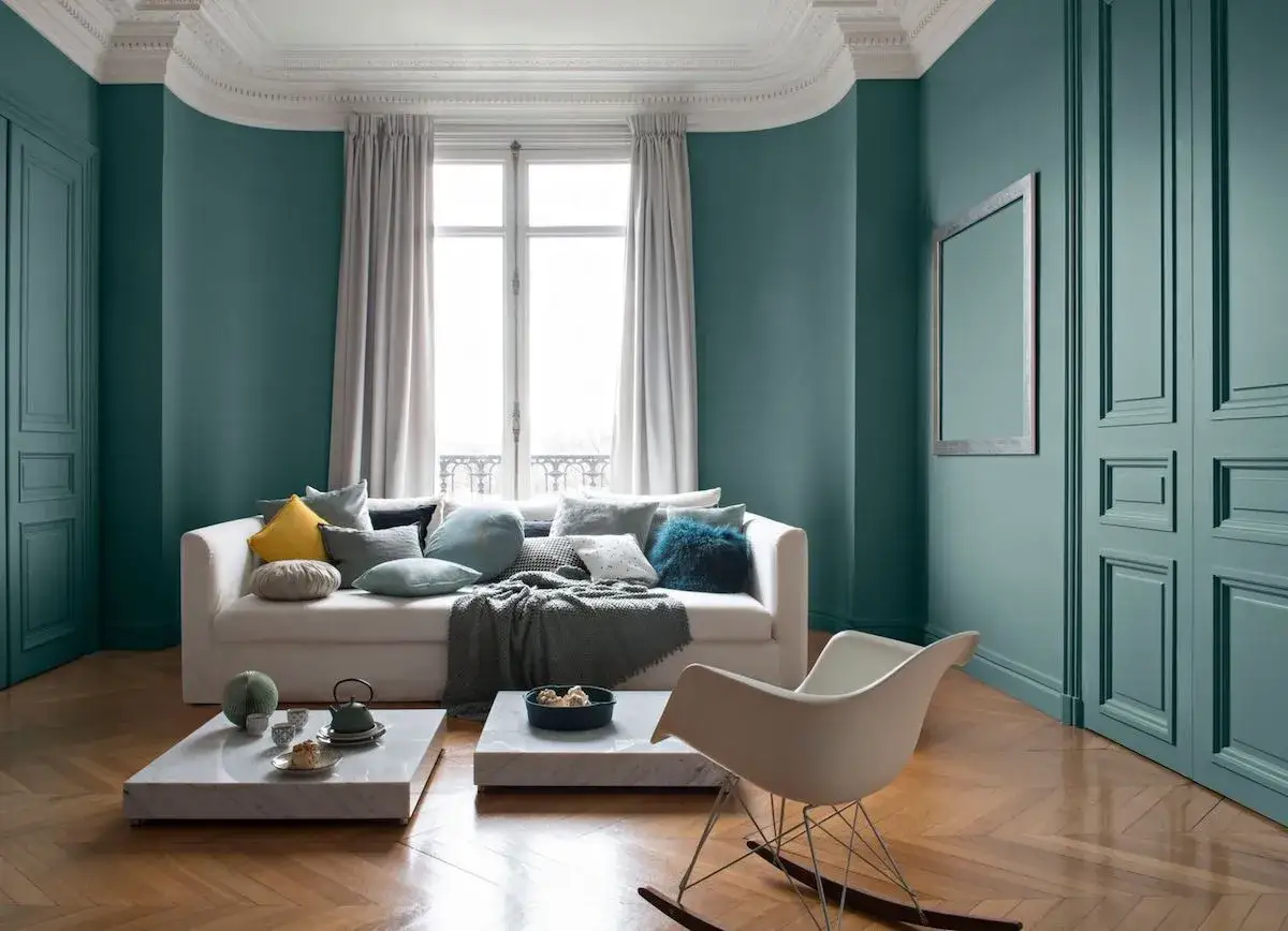 Salon moderne aux murs bleu canard ou vert canard, canap&eacute; blanc, coussins vari&eacute;s, table basse en marbre et fauteuil &agrave; bascule.