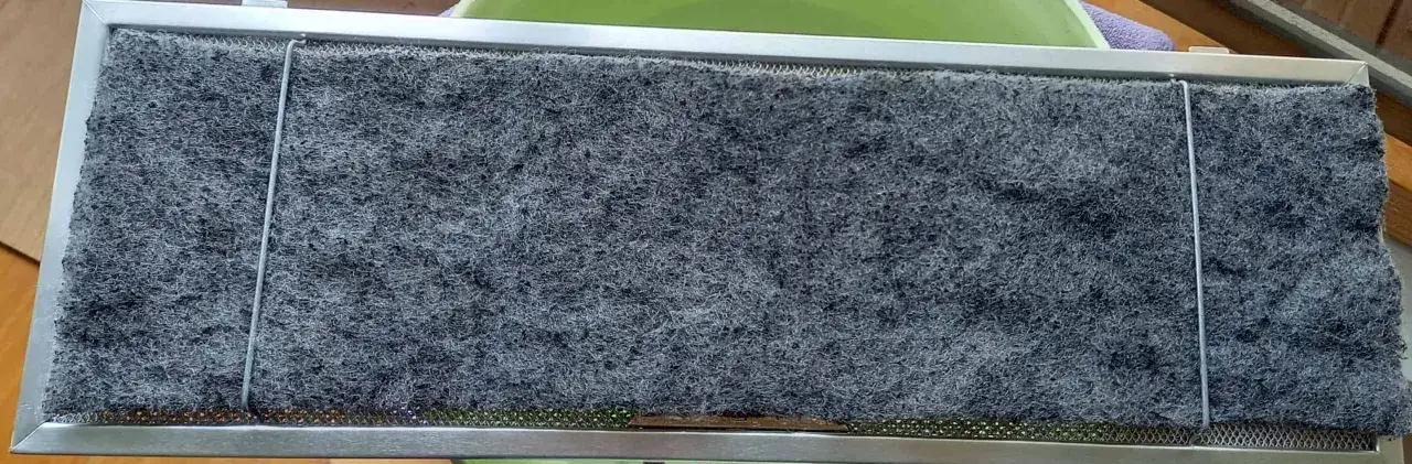 Filtre &agrave; charbon de hotte aspirante, pr&ecirc;t &agrave; nettoyer. Il est compos&eacute; d'une grille m&eacute;tallique et d'une &eacute;paisse couche de mat&eacute;riau filtrant gris.