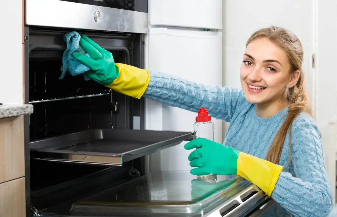 Une femme souriante, gant&eacute;e, nettoie l'int&eacute;rieur d'un four. Elle utilise un chiffon et un produit pour comment nettoyer un four catalyse.