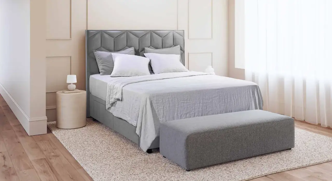 Lit de dimension queen size, avec t&ecirc;te de lit capitonn&eacute;e grise, draps blancs et banc assorti.