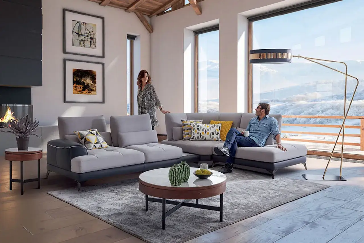 Un couple profite d'un moment de détente dans un salon moderne avec un grand canapé au milieu, face à une vue panoramique sur la neige.