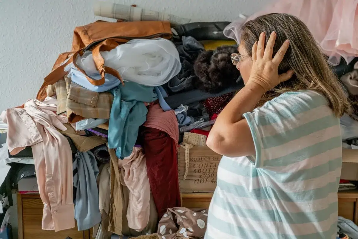 Une femme, accablée par le désordre, semble vouloir vider sa maison. Des vêtements s'entassent, créant un chaos visuel.