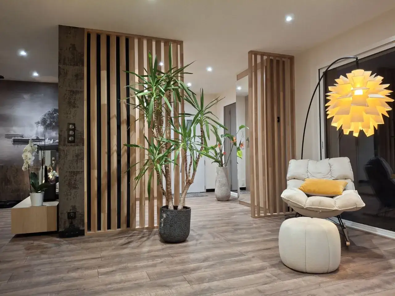 Un séparateur en bois crée une entrée dans le salon, avec une plante verte, un fauteuil confortable et une lampe design.