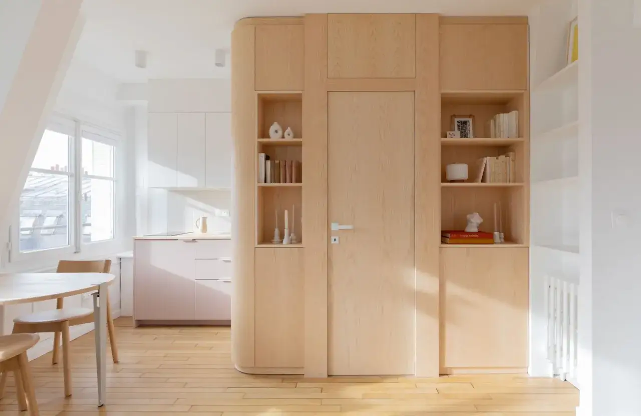 Aménagement astucieux pour optimiser petit espace : cuisine rose pâle, salle à manger bois clair, et une grande bibliothèque intégrée autour d'une porte.