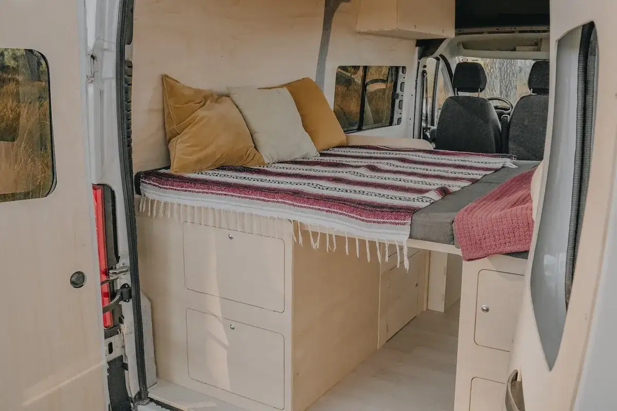 Am&eacute;nagement int&eacute;rieur d'un van avec un lit confortable, des coussins douillets et des rangements pratiques. Ce lit 1 place est parfait pour vos aventures.
