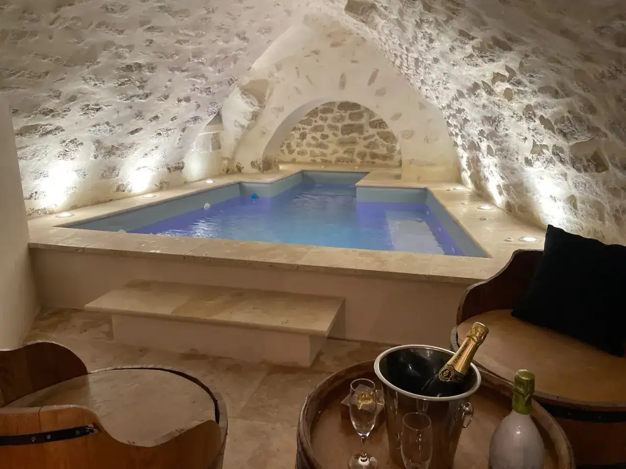 Un havre de paix souterrain : un spa dans une cave voûtée en pierre, avec une piscine illuminée et une bouteille de champagne prête à être dégustée.