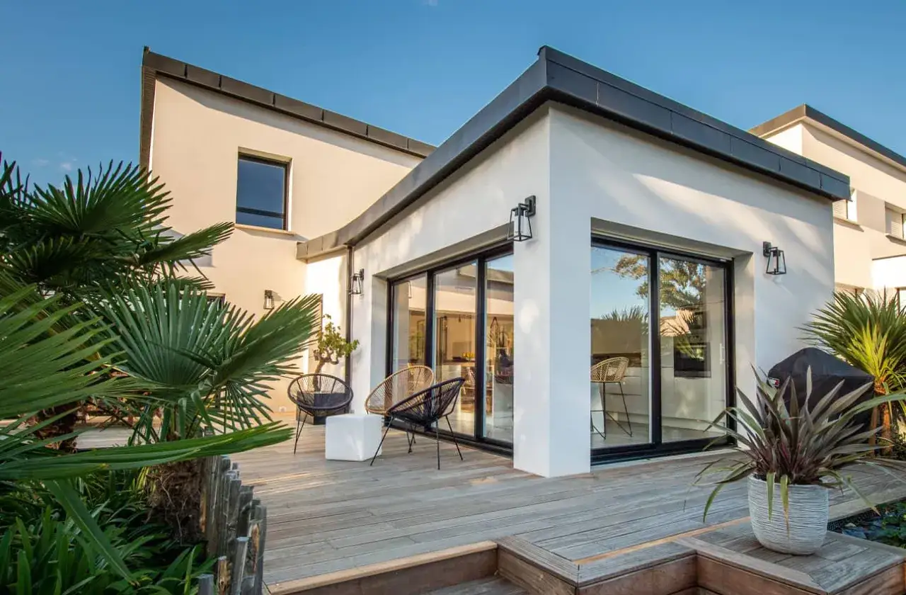 Belle extension maison avec baie vitr&eacute;e donnant sur une terrasse en bois. Palmiers et mobilier d'ext&eacute;rieur invitent &agrave; la d&eacute;tente.