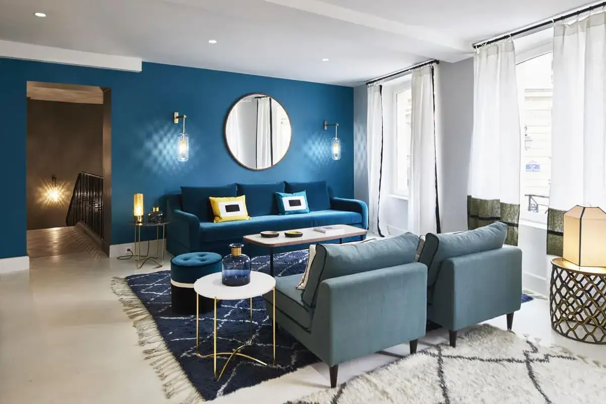 Salon moderne avec mur bleu canard, canapé velours, fauteuils et tapis assortis. Le jaune des coussins apporte une touche de chaleur.
