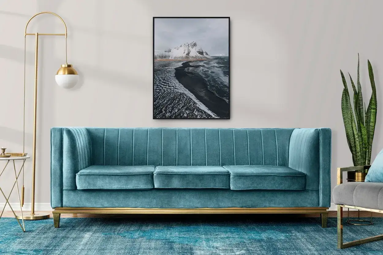 Canap&eacute; velours bleu canard, tapis assorti, et un tableau de paysage montagneux. Une lampe dor&eacute;e et une plante verte compl&egrave;tent cette d&eacute;co bleu canard et bois.