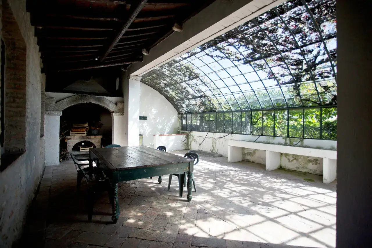 Aménagement maison en longueur : table et chaises sous une verrière ombragée, avec un four à pain en pierre.