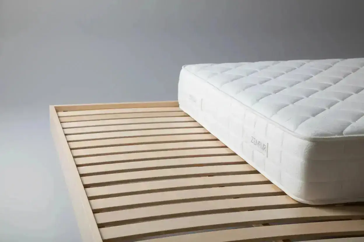 Un matelas trop grand pour sommier, ses lattes de bois en premier plan, puis le matelas blanc matelass&eacute;.