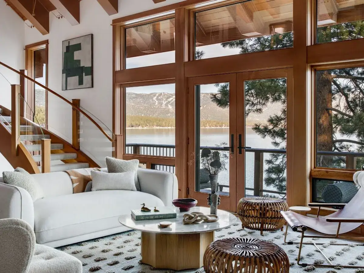 Un salon de chalet intérieur lumineux avec vue sur un lac et des montagnes. Un canapé blanc confortable, des poufs en rotin et un escalier en bois créent une ambiance chaleureuse.