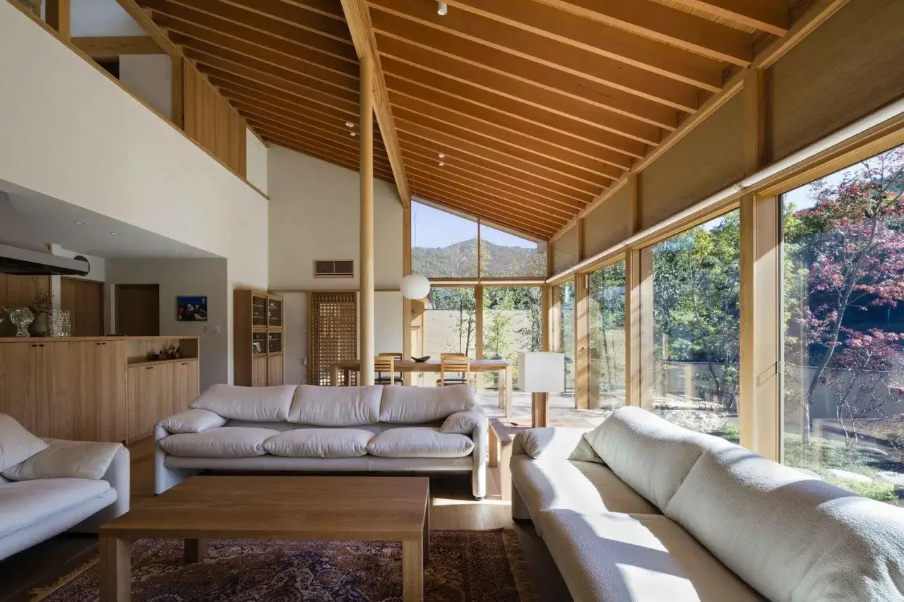 Salon lumineux d'une maison style japonais en France, avec canap&eacute;s blancs, bois apparent et grandes baies vitr&eacute;es donnant sur un jardin luxuriant.
