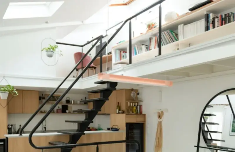 Aménagement mezzanine ouverte avec escalier métallique, cuisine en bois, étagères remplies de livres et plantes suspendues.